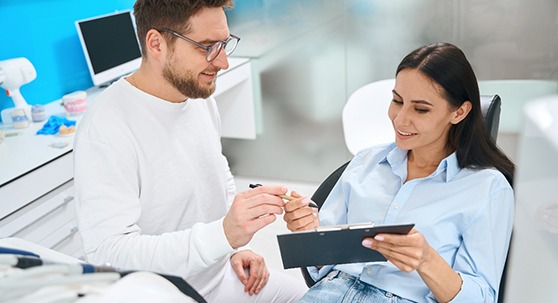 Man in white handing woman in dental chair forms on clipboard